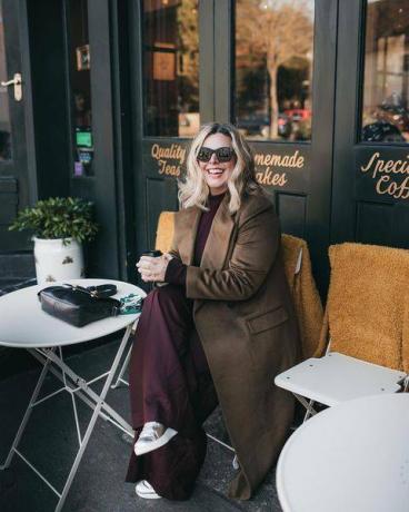 Erica Davies w czekoladowobrązowym płaszczu i brązowych spodniach siedzi przed kawiarnią