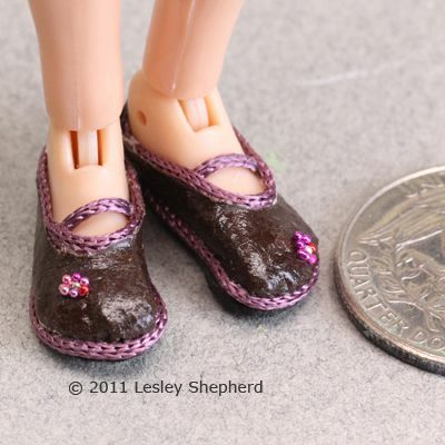 Um par de minúsculos sapatos de boneca estilo Mary Jane com cerca de 25 centavos para mostrar o tamanho.