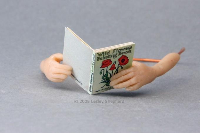 Mãos de boneca em miniatura em escala 1:12, esculpidas em argila de polímero com dedos e polegares detalhados, seguram um livro de casa de boneca