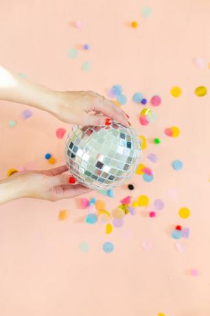 Händer som håller en DIY -diskokula på en rosa bakgrund med konfetti